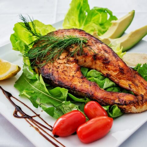 Delicious grilled salmon steak served with avocado, lettuce, cherry tomatoes, and lemon slice on a white plate.
